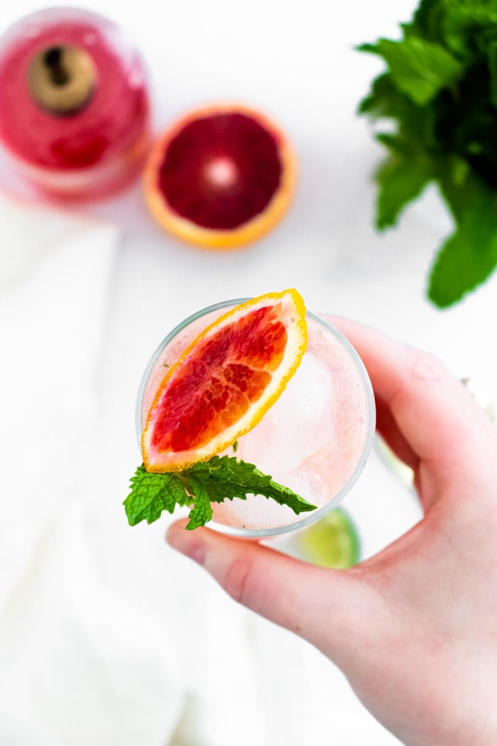 top down shot of a hand holding up Tall glass filled with ice and a blood orange mojito and blood orange slice and mint on the top