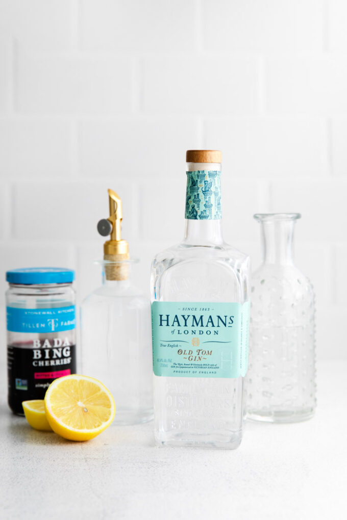 ingredients in front of a white background for tom collins: gin, lemon, cherries, simple syrup, and club soda