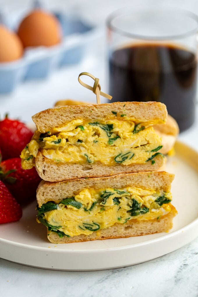 english muffin sandwich with spinach scrambled eggs topped with cheese in the middle. strawberries and coffee are in the background