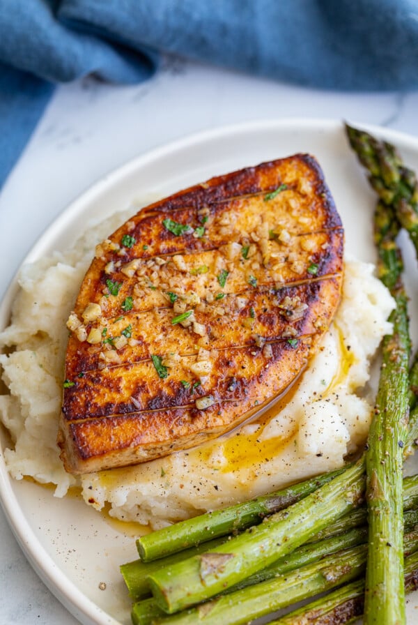 garlic butter tofu that loks a lot like a chicken breasts sitting on top of mashed potatoes with roasted asparagus on he side of the plate