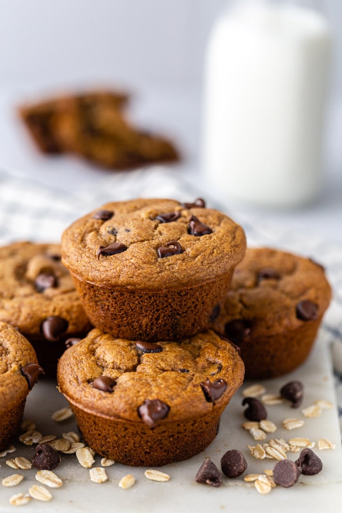 stack of chocolate oat flour muffins with milk and another muffin in the background. Loose chocolate chips and oats around the scene