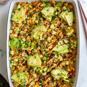 vegetarian sushi bake inside of a casserole dish that's topped with heaps of avocado, sesame seeds and drizzled with red sriracha, orange spicy mayo, and kewpie mayo. It's also sprinkled with chopped green onion